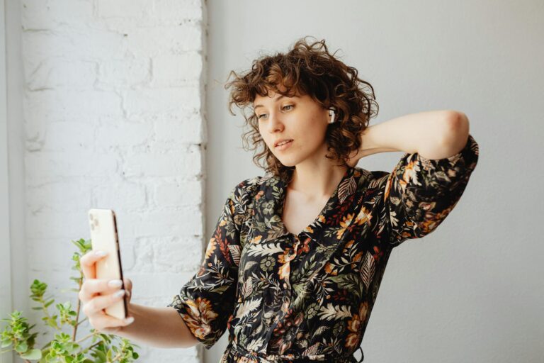Woman with curly hair taking selfie in floral dress, using a smartphone and wireless earbuds.