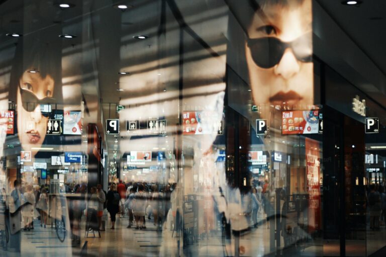 Reflections of shoppers and fashion posters in a modern mall with bright lights.