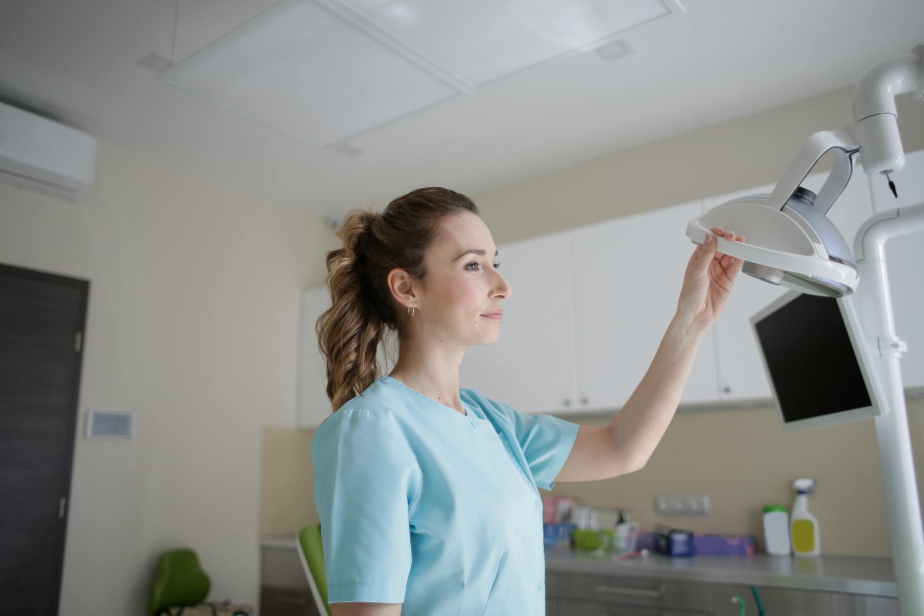 Thoughtful female medic in blue uniform touching medical lamp while standing near computer screen in clinic and looking away in daylight