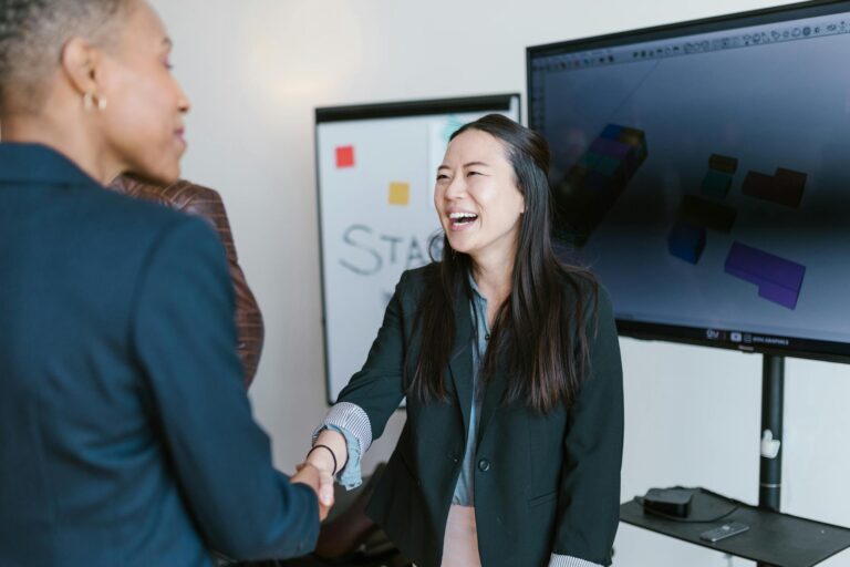 Two businesswomen shake hands while smiling in a modern office setting, emphasizing teamwork and collaboration.