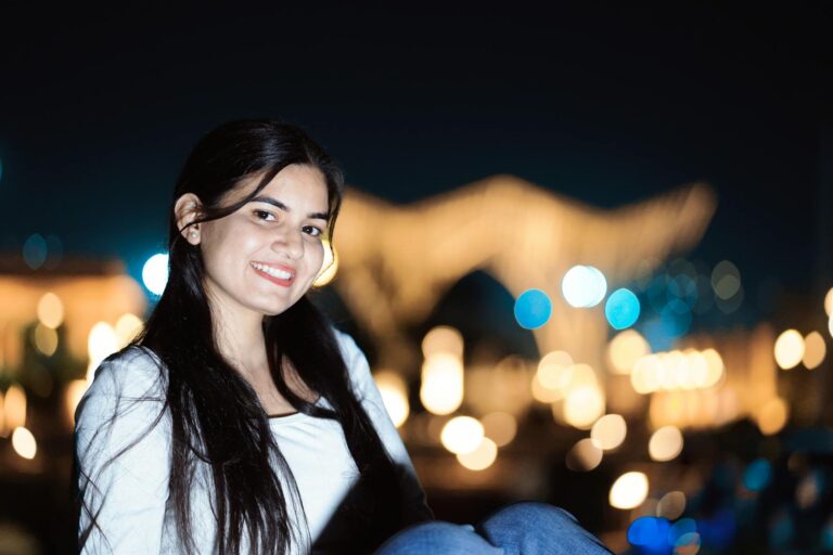 A young woman smiling against a blurry city lights backdrop at night.